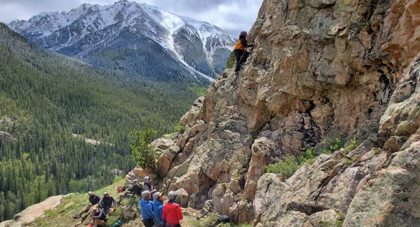 With a snowcapped mountain in the background, a rock climber ascends a rock face while a group of people watch from below.